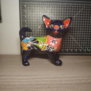 Pode incluir: Uma estatueta de chihuahua de cerâmica em posição de pé. O cão é principalmente azul escuro com orelhas vermelho-alaranjadas e um peito rosa e com padrão floral. As costas do cão são decoradas com desenhos florais coloridos. A estatueta tem um acabamento brilhante.