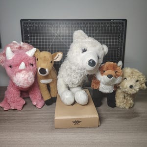 May include: A selection of soft toy animals: a pink triceratops, a brown deer, a white polar bear, a red fox, and a tan poodle. The toys are displayed on a wooden surface, with a cutting mat in the background.