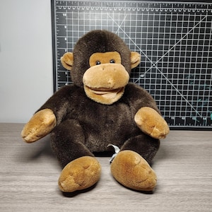 May include: A plush monkey toy with brown fur, tan face, hands, and feet. The stuffed animal has a smiling expression and is sitting upright. The background includes a black cutting mat and a wooden surface.