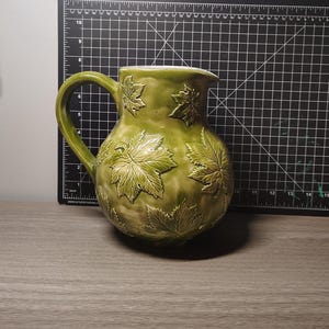 May include: A green ceramic jug with a large handle and a spout. The jug is decorated with raised leaf designs. The jug is sitting on a wooden surface.