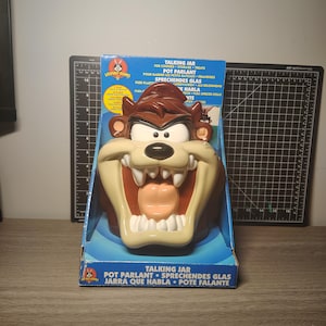 Könnte beinhalten: Ein Looney Tunes Taz Sprechglas für Kekse, Aufbewahrung und Leckereien. Das Glas hat die Form von Taz's Kopf, mit braunem Oberteil, beigem Gesicht und offenem Mund. Die Verpackung ist blau mit Text in mehreren Sprachen.