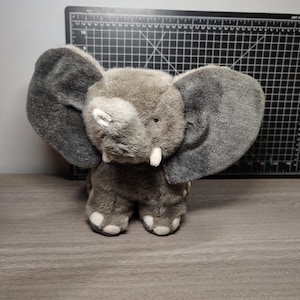 Può includere: Un peluche a forma di elefante con grandi orecchie grigie e un corpo grigio. L'elefante ha zanne bianche e accenti bianchi sui piedi. Il giocattolo è realizzato in materiale morbido e soffice ed è progettato per assomigliare a un elefantino.