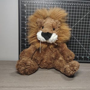 May include: A plush lion toy with a brown body and a fluffy, light brown mane. The lion has a black nose, white muzzle, and is sitting on a wooden surface. The toy is approximately 10 inches tall.