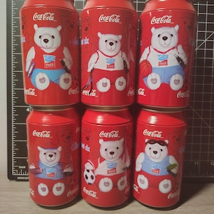 Può includere: Sei lattine rosse da collezione Coca-Cola con un orso polare in vari abiti sportivi. Ogni lattina ha il logo Coca-Cola e illustrazioni di orsi. Le lattine sono alte circa 13 cm.