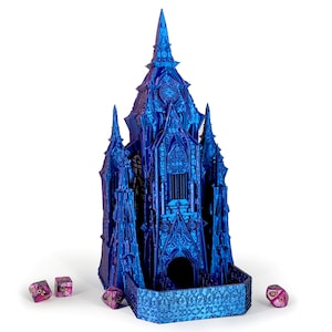May include: A blue and purple iridescent gothic castle-shaped dice tower with a detailed design. The tower has a small opening at the bottom for dice to roll out. Four pink dice are scattered on a white surface.