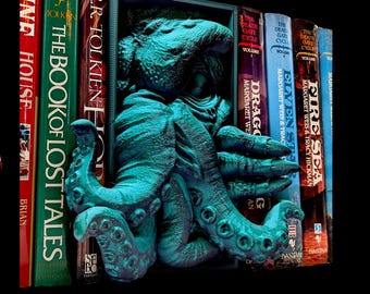 May include: A teal-colored, 3D-printed bookend featuring an octopus design. The tentacles wrap around a row of books, including titles like "The Book of Lost Tales" and "Fire Sea." The bookend is set against a black background.
