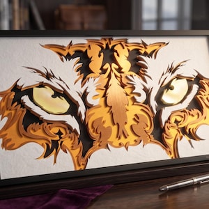 Op de afbeelding: Een rechthoekig ingelijst wandkunstwerk met een meerlaags, lasergesneden papierontwerp van een tijgergezicht. Het kunstwerk toont intense gele ogen, ingewikkelde oranje en gouden vachtpatronen en diepe zwarte schaduwen in een zwarte lijst.
