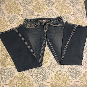 Puede incluir: Vaqueros de mezclilla azul oscuro con dise&ntilde;o de pernera acampanada. Los vaqueros presentan costuras blancas en las costuras y los bolsillos. Los vaqueros est&aacute;n colocados sobre una tela estampada.