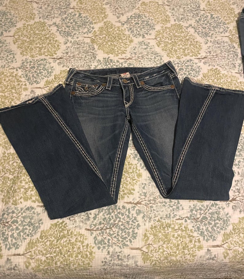 Puede incluir: Vaqueros de mezclilla azul oscuro con dise&ntilde;o de pernera acampanada. Los vaqueros presentan costuras blancas en las costuras y los bolsillos. Los vaqueros est&aacute;n colocados sobre una tela estampada.