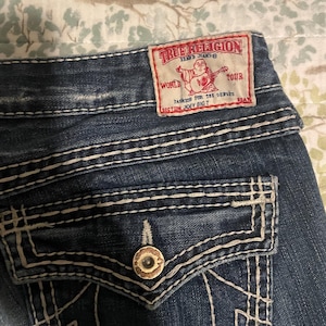 Puede incluir: Primer plano de unos vaqueros de mezclilla azules con costuras blancas y un bolsillo trasero. Es visible una etiqueta True Religion roja y blanca. La etiqueta presenta un gr&aacute;fico de una persona tocando la guitarra y el texto "WORLD TOUR".