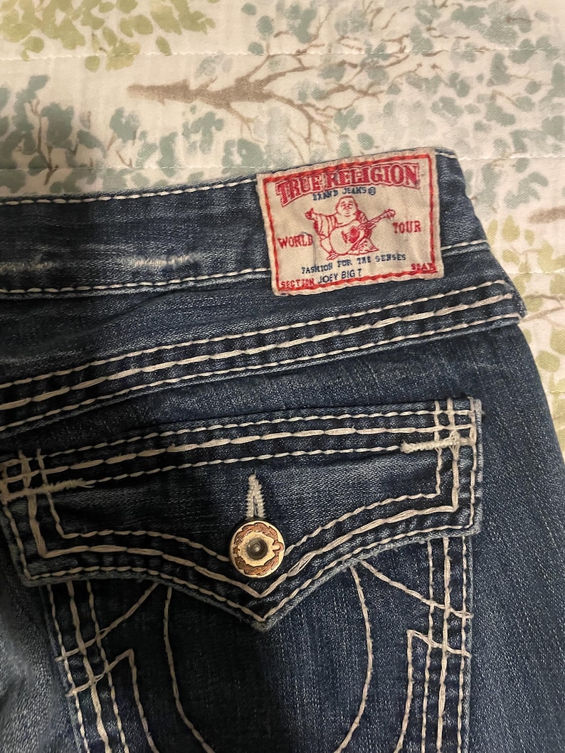 Puede incluir: Primer plano de unos vaqueros de mezclilla azules con costuras blancas y un bolsillo trasero. Es visible una etiqueta True Religion roja y blanca. La etiqueta presenta un gr&aacute;fico de una persona tocando la guitarra y el texto "WORLD TOUR".