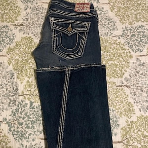 Puede incluir: Vaqueros de mezclilla azul oscuro con costuras blancas y un dise&ntilde;o cl&aacute;sico de cinco bolsillos. Cuenta con cierre de bot&oacute;n y cremallera, y un parche de la marca True Religion. Los vaqueros est&aacute;n doblados por la mitad.