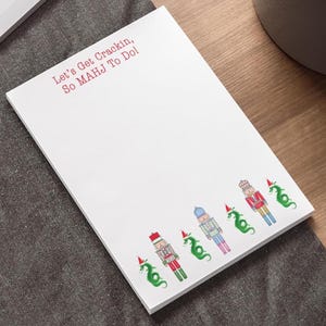 May include: White notepad with the text "Let's Get Crackin, So MAHJ To Do!" in red. The bottom of the notepad features illustrations of nutcrackers and green dragons wearing Santa hats. The notepad is on a gray surface.