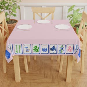 May include: A light pink tablecloth with a border of illustrated tiles featuring various designs, including a green dragon, blue flowers, and a hummingbird. The table is set with white plates and silverware, and surrounded by wooden chairs.