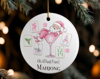 Könnte beinhalten: Ein weißer, runder Weihnachtsschmuck mit einem goldenen Faden. Er zeigt einen rosa Weihnachtsmann, der sich in einem rosa Cocktailglas entspannt, umgeben von Mahjong-Steinen. Der Text "Oh What Fun! Mahjong" ist darunter gedruckt.