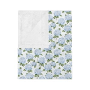 Puede incluir: Una manta de forro polar blanca y suave con un estampado floral azul y verde con hortensias. La manta es reversible con un lado de forro polar blanco y un lado floral.