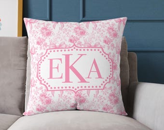 Può includere: Un cuscino quadrato con sfondo bianco e motivo floreale rosa. Il cuscino presenta un design incorniciato in rosa con le lettere "EKA" in rosa. Il cuscino è su un divano grigio.