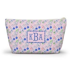 May include: Pink cosmetic bag with a pattern of Mahjong tiles and blue dice. The bag has a blue rectangular frame with the letters "KBA" in a blue script font. The zip is gold.