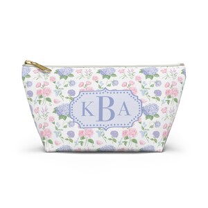 Puede incluir: Un neceser blanco con un estampado floral de rosas rosas y hortensias azules. La bolsa presenta un óvalo azul claro con las iniciales "KBA" en una fuente serif. Una cremallera dorada asegura la parte superior.