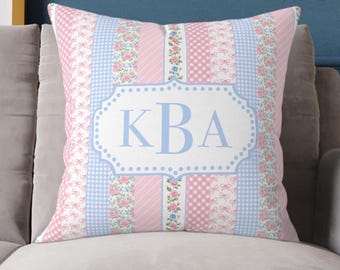 May include: Square cushion with a patchwork design in shades of pink, white, and blue. The cushion has a central white label with the light blue letters "KBA". The design includes floral, gingham, and striped patterns.