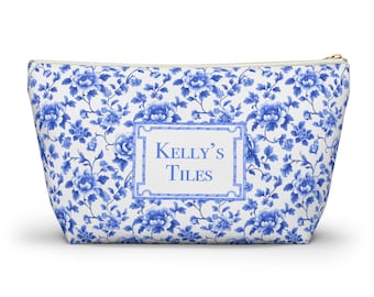 May include: A zippered pouch featuring a white background with a blue floral pattern. Centered on the front is a rectangular frame containing the text Kelly's Tiles in blue lettering. The pouch has a tapered shape with a gold-toned zipper closure.