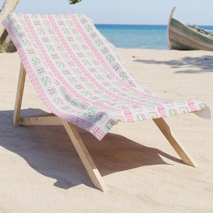 May include: A wooden beach chair with a white, pink, and green patterned beach towel. The towel features a repeating geometric design. The chair is on a sandy beach with the ocean and a boat in the background.
