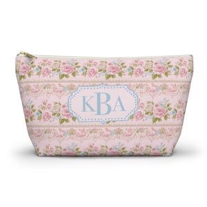 以下が含まれることがあります： ピンク色の花柄コスメポーチ、ゴールドのジッパー付き。 ピンクのバラ、緑の葉、青のアクセントの繰り返し模様が特徴です。 中央には「KBA」のイニシャルが入った水色の楕円があります。