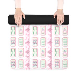 May include: A white yoga mat with a pink and green pattern of mahjong tiles. The tiles feature various symbols, including a pagoda, flowers, and bamboo.