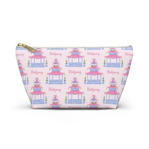 May include: A light pink zipped pouch featuring a repeating pattern of pink and blue pagodas and the word "Mahjong". The pagodas have pink roofs, blue columns, and green accents. The zip is gold.