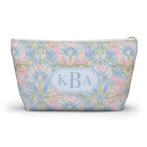 Puede incluir: Un neceser con un estampado floral en azul pastel, rosa y verde. La bolsa presenta una etiqueta rectangular azul claro con las iniciales "KBA" en una fuente azul a juego. La bolsa tiene una cremallera dorada.