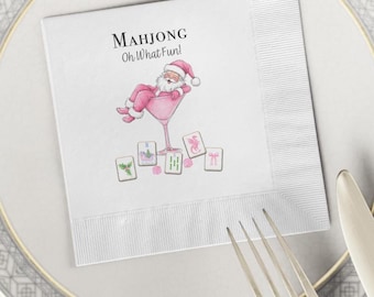 May include: White paper napkin with the text "MAHJONG Oh What Fun!" and a watercolor illustration of Santa Claus in a pink martini glass. Several Mahjong tiles are scattered around the glass. The napkin is on a white plate with a fork and knife.