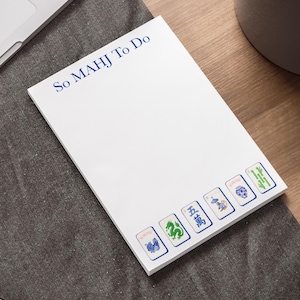May include: White notepad with the text "So MAHJ To Do" in blue. The bottom of the notepad features six Mahjong tile illustrations in blue, green, and white. The notepad is on a gray surface.