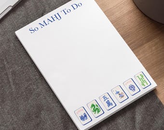 May include: White notepad with the text "So MAHJ To Do" in blue. The bottom of the notepad features six Mahjong tile illustrations in blue, green, and white. The notepad is on a gray surface.