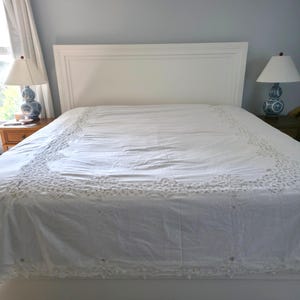 May include: A white bedspread featuring an intricate lace border with floral embroidery and cutwork details. The fabric is smooth and covers the entire bed, extending down the sides with a scalloped edge finish along the bottom hemline.