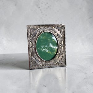 May include: A square silver-toned picture frame featuring an ornate, embossed leaf and scroll pattern surrounding a central oval opening. The frame has a metallic finish with dark recessed details and a reflective surface showing a green reflection.