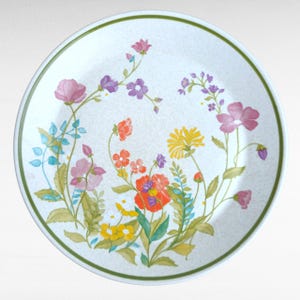 May include: Round white plate with a floral design. The plate features a green border and a speckled background. The floral pattern includes pink, purple, yellow, and red flowers with green leaves and stems.