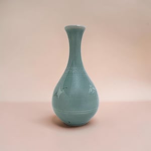 May include: A celadon-colored ceramic vase with a slender neck and a rounded body. The vase features delicate etched designs and a crane motif. The vase is approximately 8 inches tall, perfect for displaying flowers or as a decorative piece.