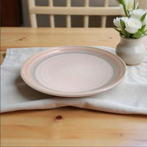 Op de afbeelding: Een ronde keramische plaat met een lichtroze midden en concentrische ringen van roze, grijs en donkerder roze rond de rand. De plaat ligt op een wit linnen servet op een houten tafel. Rechts staat een kleine vaas met witte bloemen.