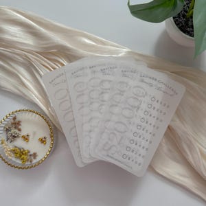 May include: A stack of translucent white savings challenge cards with the text "Savings Challenge Twenty Thousand". Each card has circles for tracking savings amounts. A decorative candle with dried flowers sits nearby.