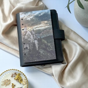 May include: A black leather-textured planner with a six-hole punched insert featuring a detailed photographic print of a castle nestled in a mountainous landscape under a cloudy sky. The insert includes the small printed text MIKALACREATIONSCO.