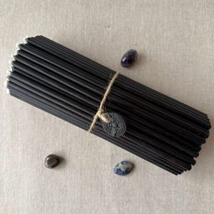 May include: A bundle of black incense sticks tied with twine and a small, round tag. The incense is accompanied by three small, polished gemstones: amethyst, labradorite, and sodalite. The background is a neutral, textured fabric.