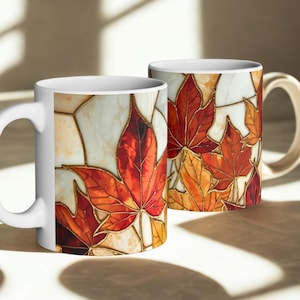 May include: Two white ceramic mugs with a stained glass design of autumn leaves in shades of red, orange, and yellow. The mugs have a white handle and the text "Stained glass design on a ceramic mug" at the bottom.