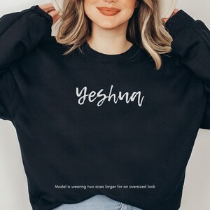 May include: Black sweatshirt with the word "Yeshua" in white script across the chest. The sweatshirt is a solid black color and has a crew neck. The text "Model is wearing two sizes larger for an oversized look" is at the bottom.