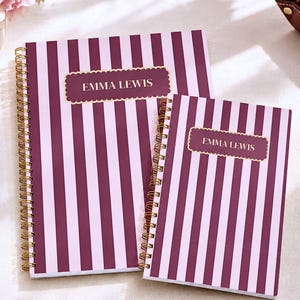 May include: Two spiral-bound notebooks featuring a vertical striped pattern in dark maroon and light pink. Each notebook has a central rectangular label with a scalloped gold border containing the white printed text EMMA LEWIS.