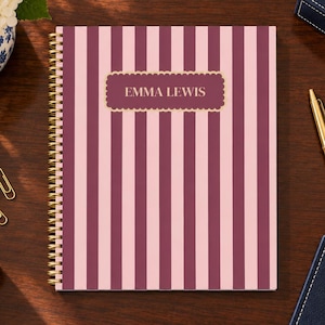 May include: A spiral-bound notebook featuring vertical stripes in alternating shades of light pink and dark maroon. A central rectangular label with a scalloped gold border displays the name EMMA LEWIS in gold capital letters.