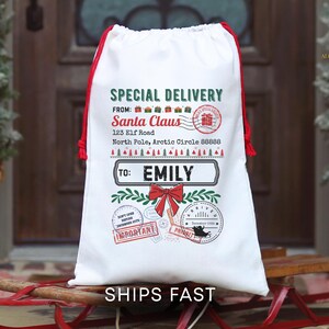 Könnte beinhalten: Ein weißer Santa Claus Geschenkebeutel mit rotem Kordelzug und Akzenten. Die Tasche trägt den Text "Special Delivery" und "From Santa Claus" mit einer Adresse und "To: Emily". Dekorative Stempel und eine rote Schleife vervollständigen das Design. Der Text "Ships Fast" steht unten.
