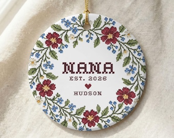May include: A round white ceramic ornament featuring a cross-stitch style floral wreath with red and blue flowers and green leaves. The text reads NANA EST. 2026 with a small red heart above the name HUDSON in dark maroon cross-stitch lettering.