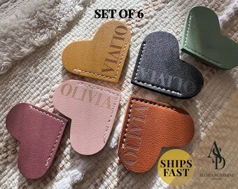 May include: Set of six heart-shaped leather bookmarks in various colors: mustard yellow, black, olive green, rose, light pink, and brown. Each bookmark has the name "OLIVIA" and white stitching. The text "SET OF 6" is at the top.
