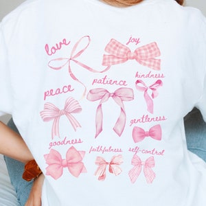 May include: White t-shirt with a watercolour design of pink bows and words such as "love", "joy", and "kindness". The bows are in various styles, including gingham and ribbon designs. The words are in a delicate, handwritten font.