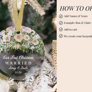 K&ouml;nnte beinhalten: Ein runder, durchsichtiger Acryl-Weihnachtsbaumschmuck mit einem Blumenarrangement aus wei&szlig;en und blassrosa Rosen sowie gr&uuml;nem Blattwerk. Der Text lautet: Our First Christmas MARRIED Doug & Linda 2026. Ein goldenes Band ist oben zum Aufh&auml;ngen befestigt.